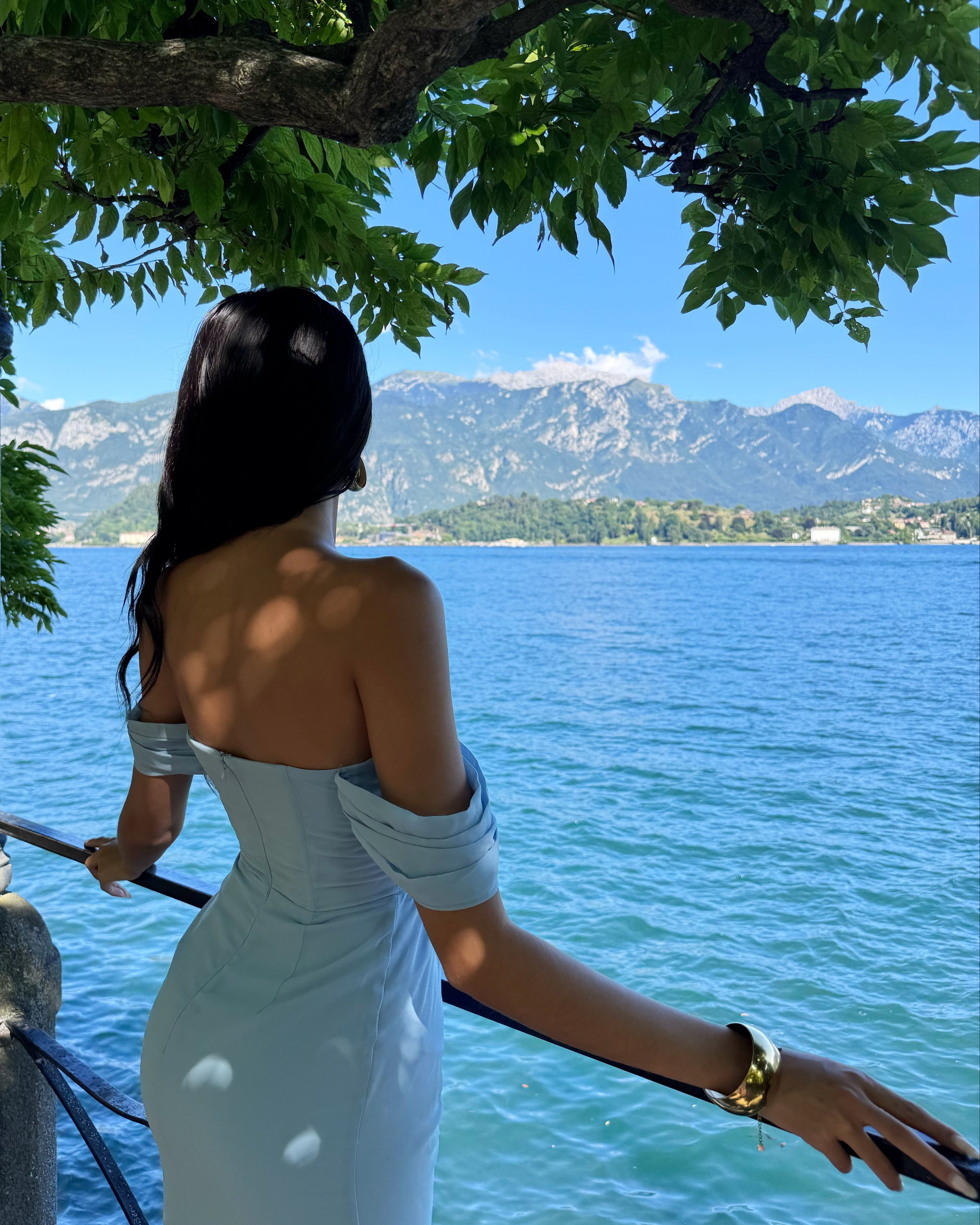 Woman in a light blue dress standing by a railing overlooking a lake with mountains in the background