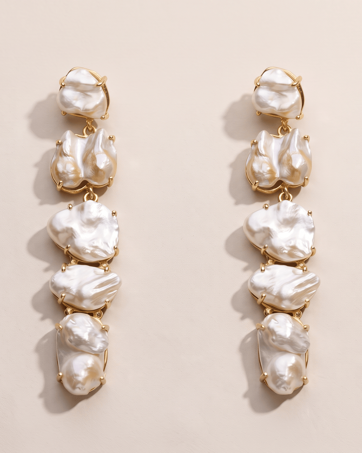 A pair of gold drop earrings featuring five vertical clusters of irregular, lustrous white baroque pearls, displayed on a warm beige background.