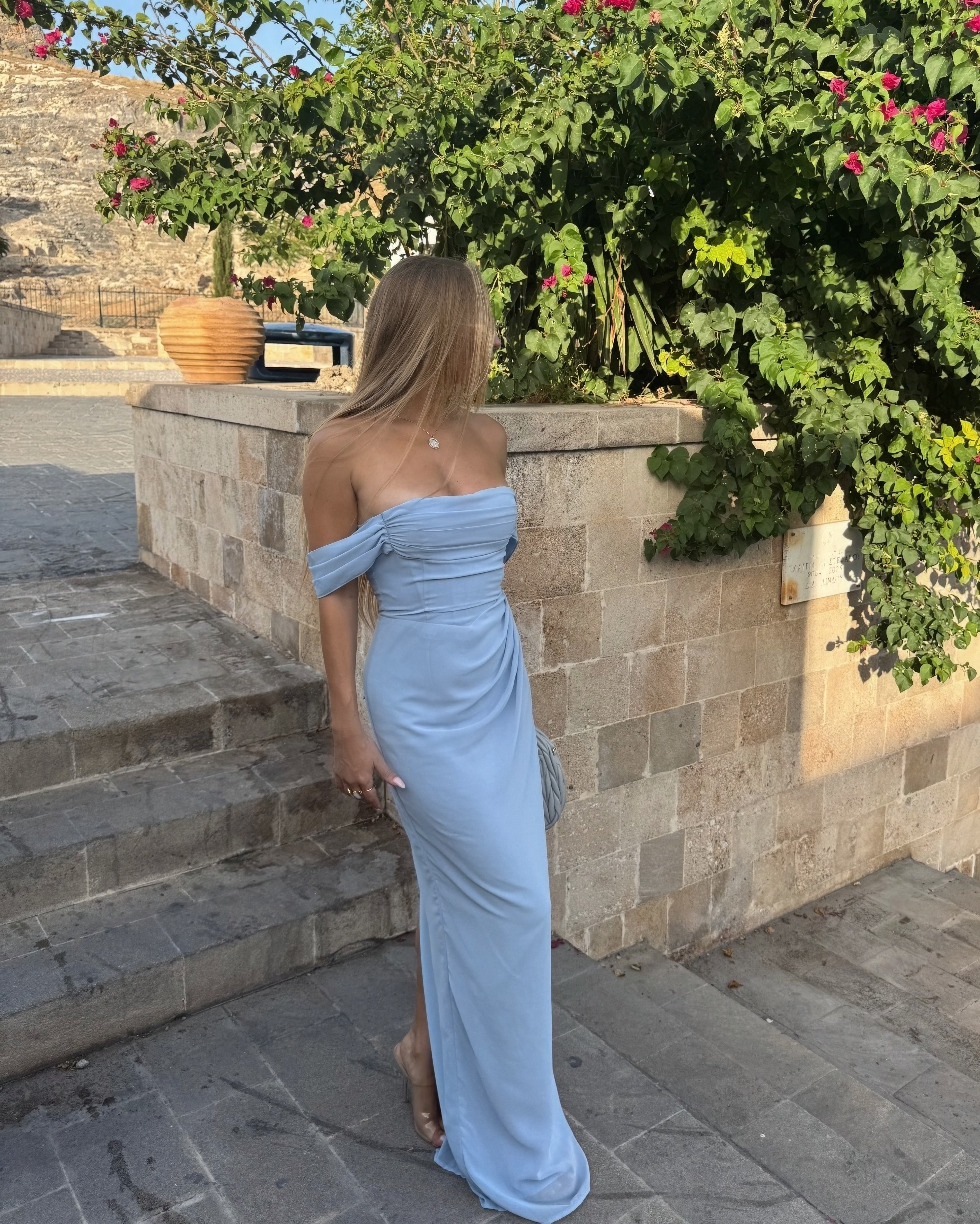 Woman in a light blue off-shoulder dress standing outdoors near stone steps and greenery.