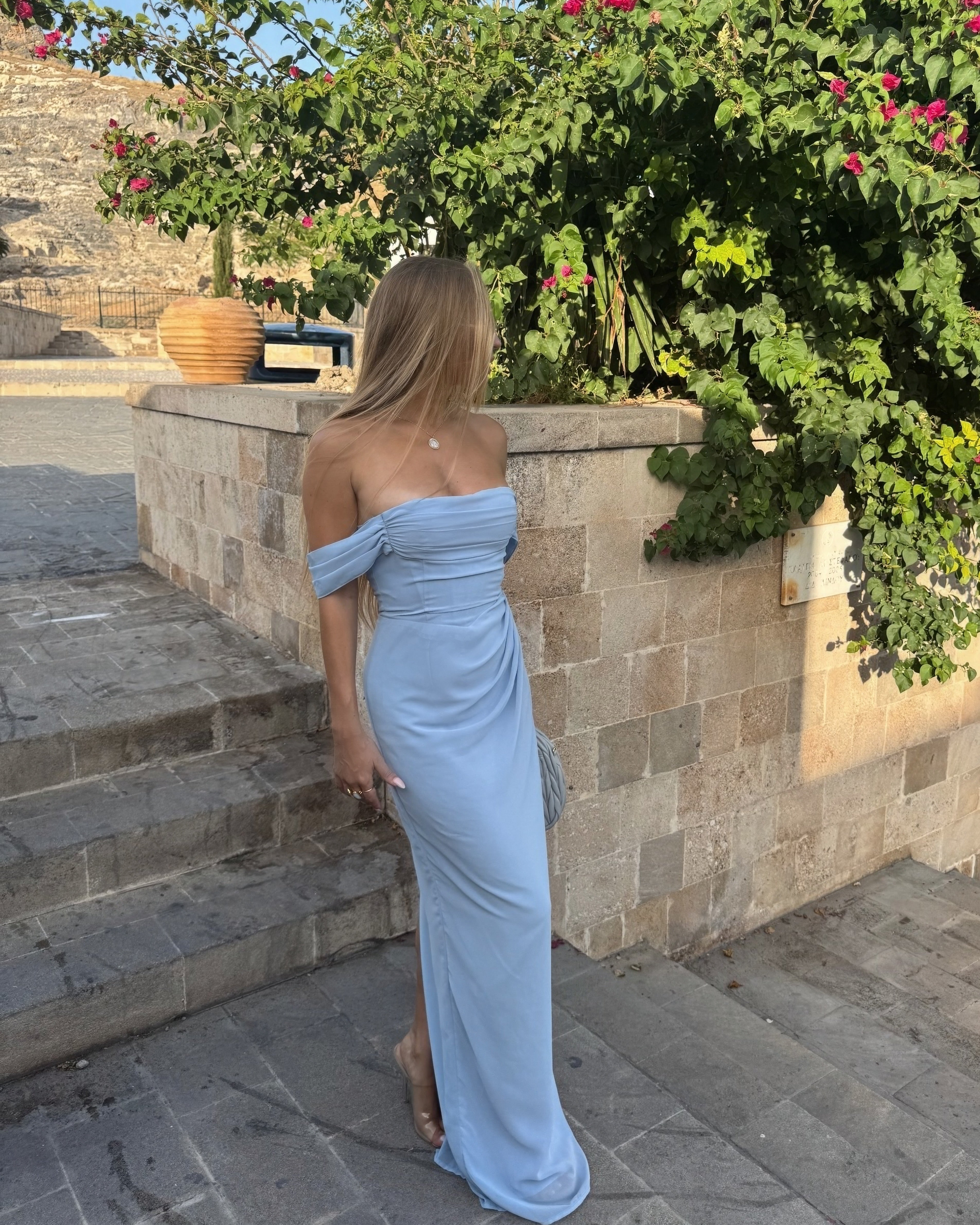 Woman in a light blue off-shoulder dress standing outdoors near stone steps and greenery.