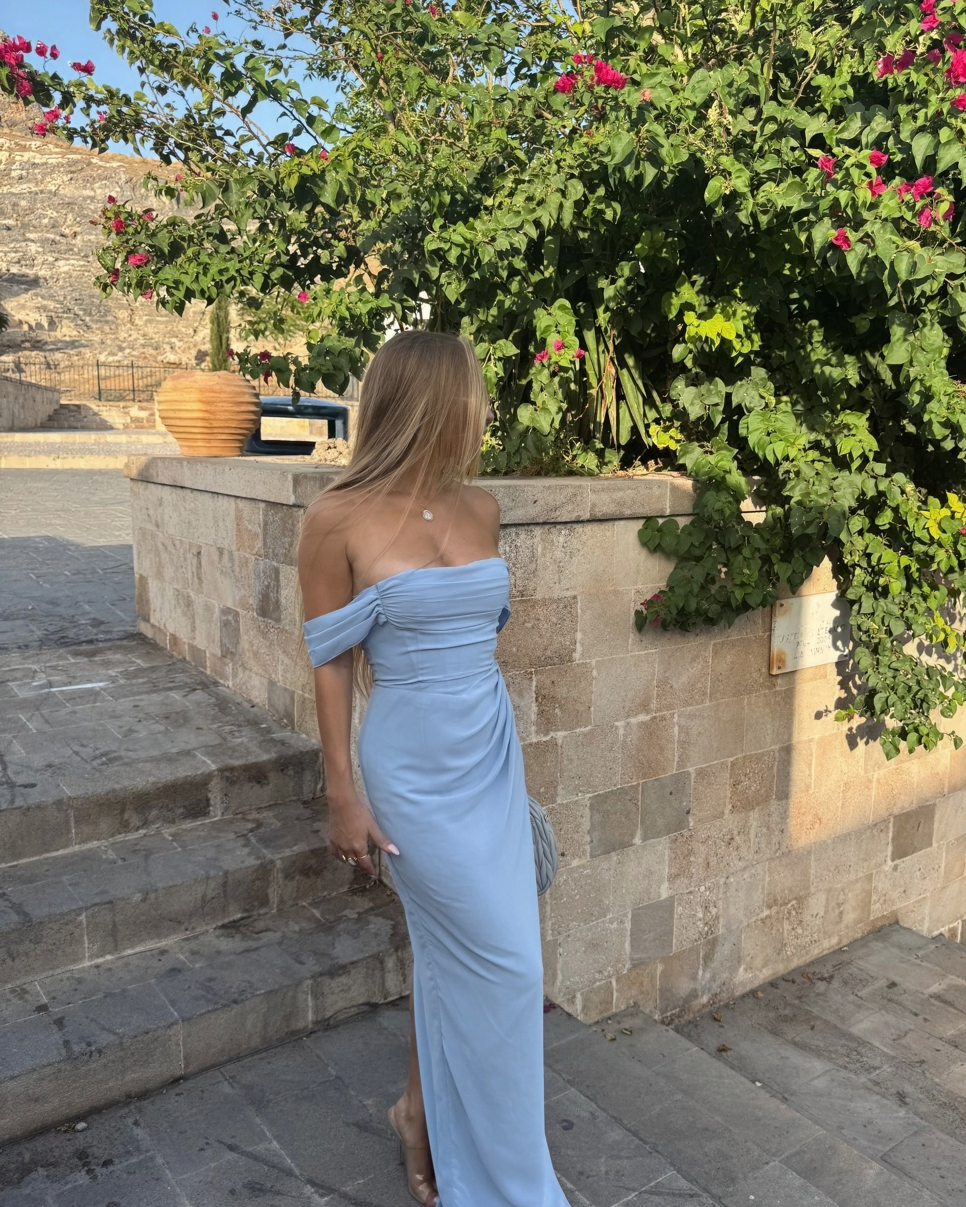 Woman in a light blue off-shoulder dress standing outdoors near stone steps and greenery.