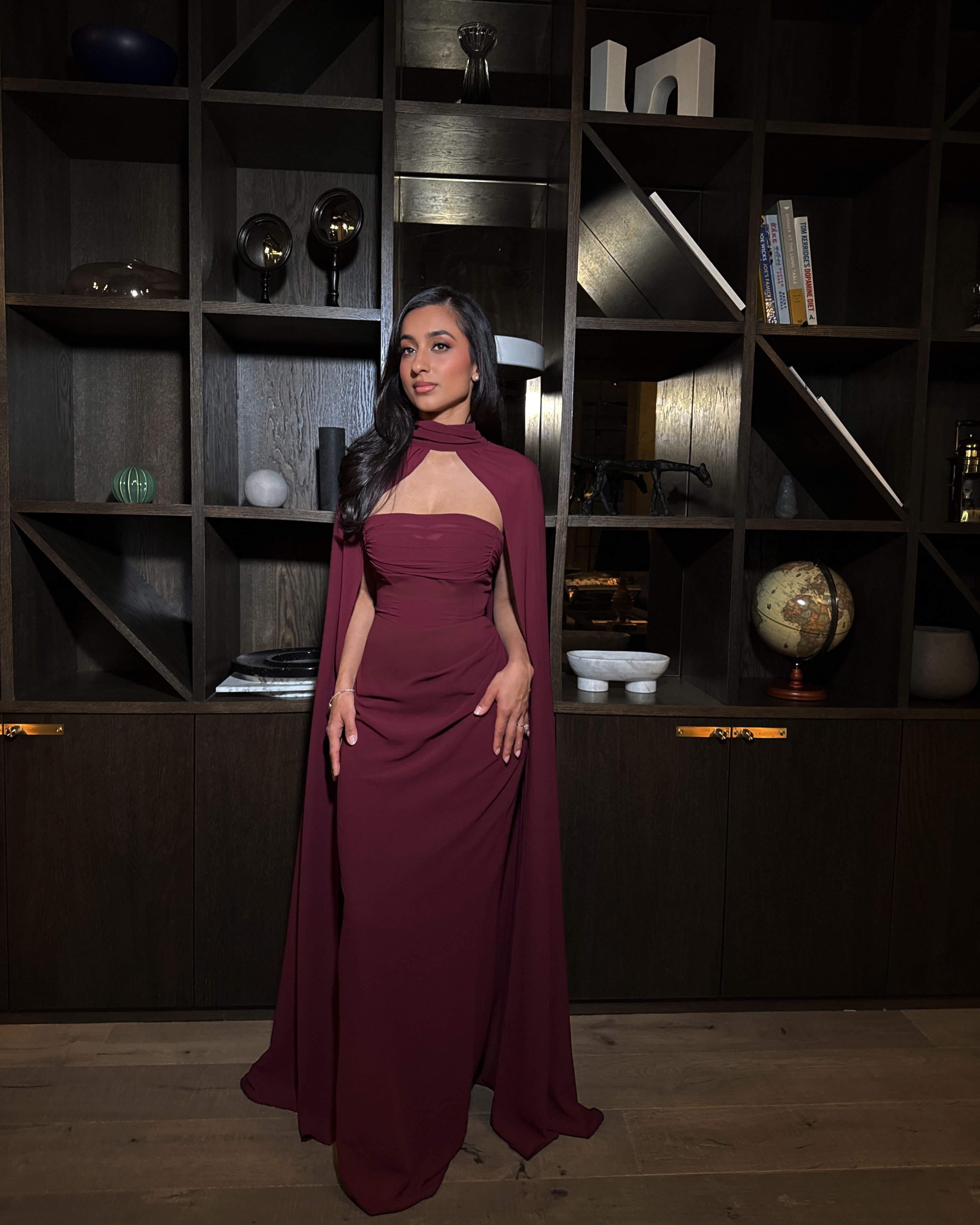 Woman in a burgundy dress standing in a modern interior setting with shelves and decor.