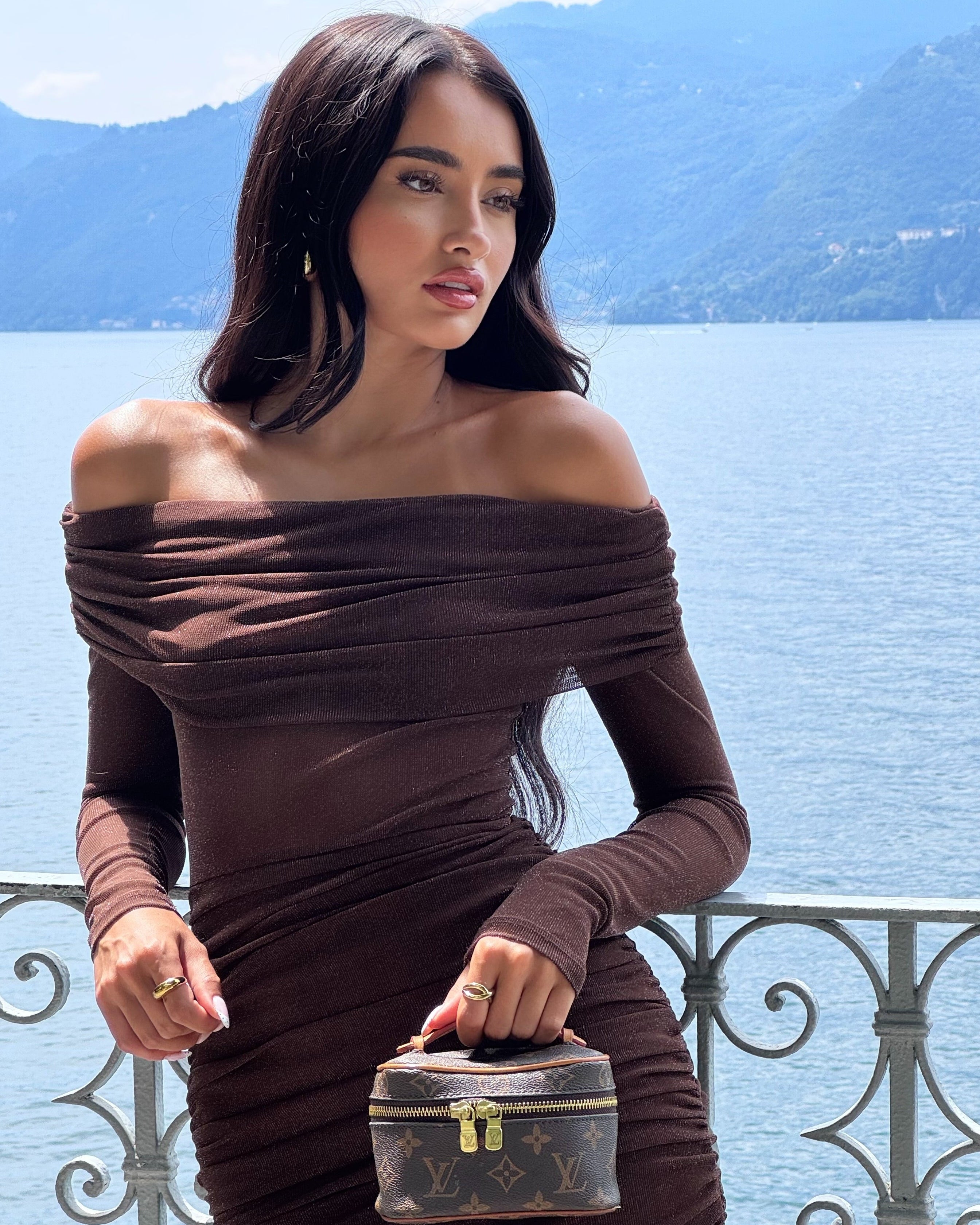 Woman in a brown off-shoulder dress holding a small suitcase by a scenic water body.