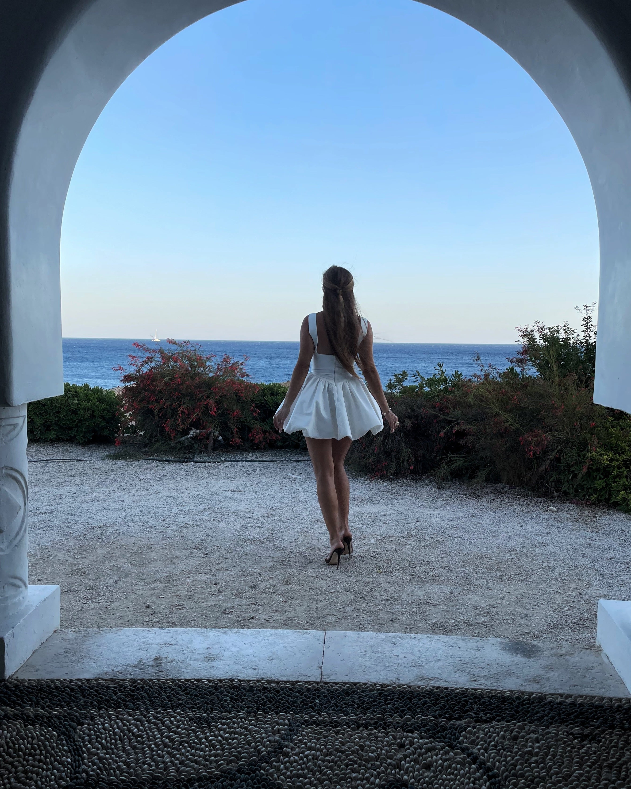 Woman in a white dress standing in front of an archway with a scenic view of the ocean.