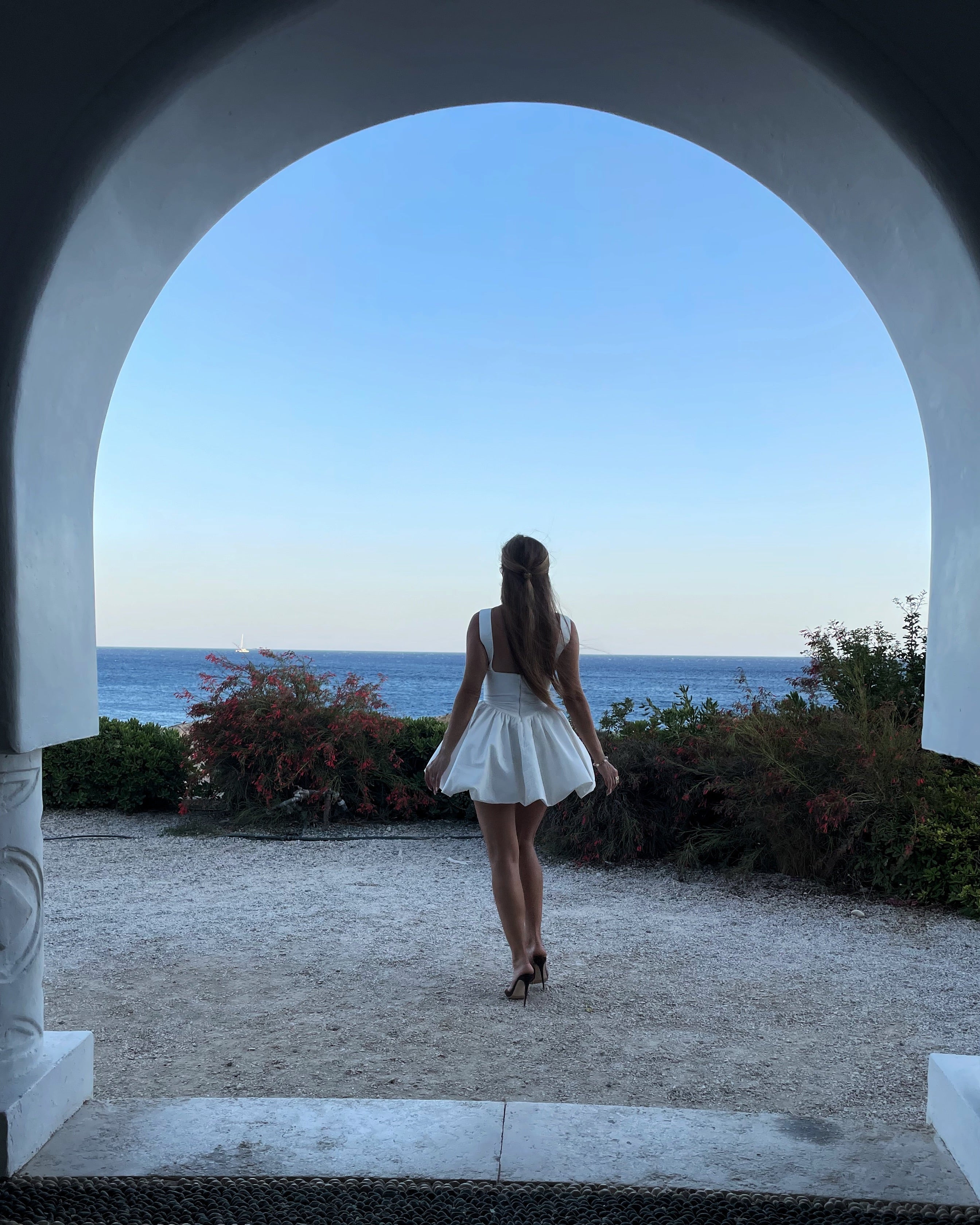 Woman in a white dress standing in front of an archway with a scenic view of the ocean.