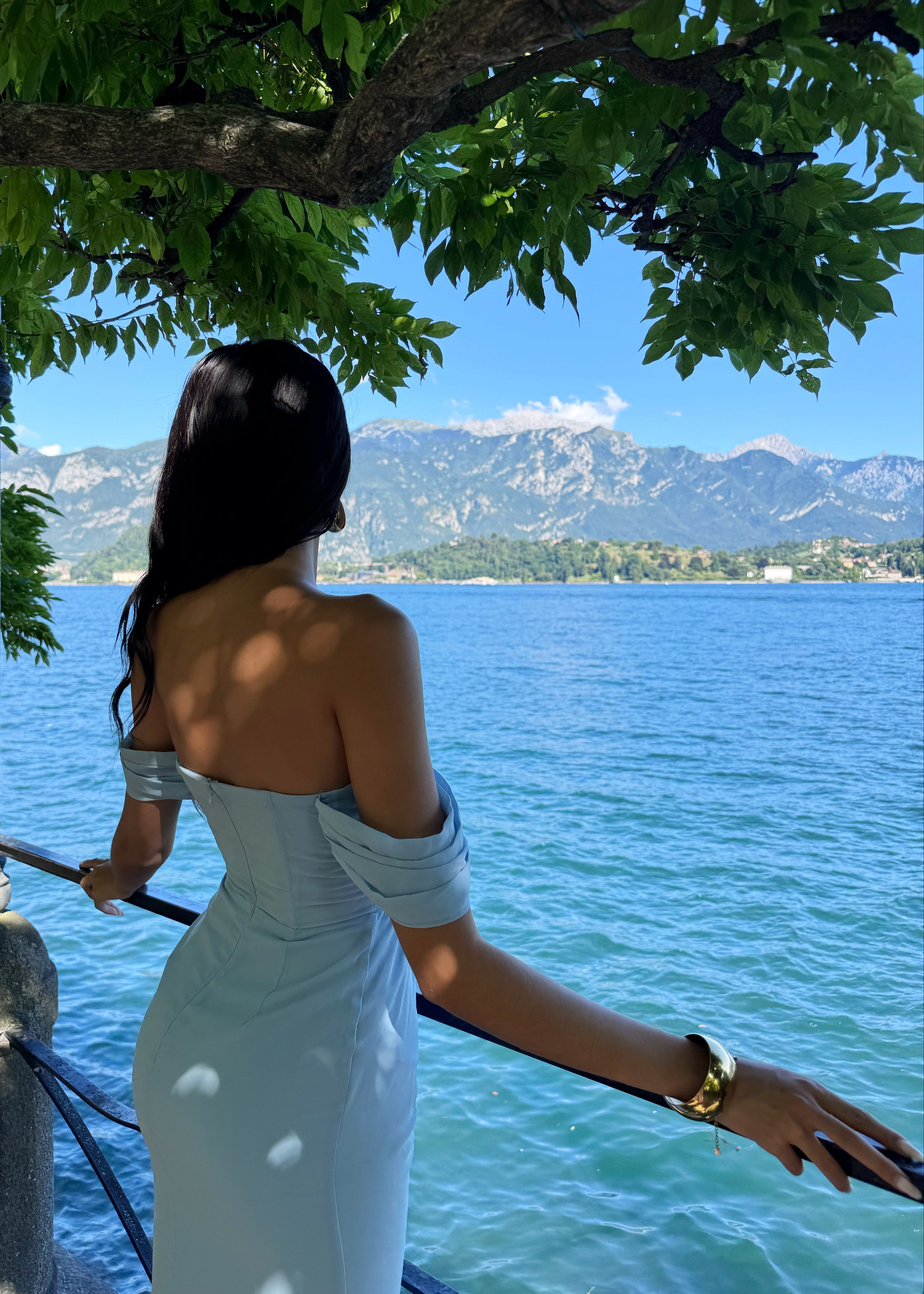 Woman in a light blue dress standing by a railing overlooking a lake with mountains in the background
