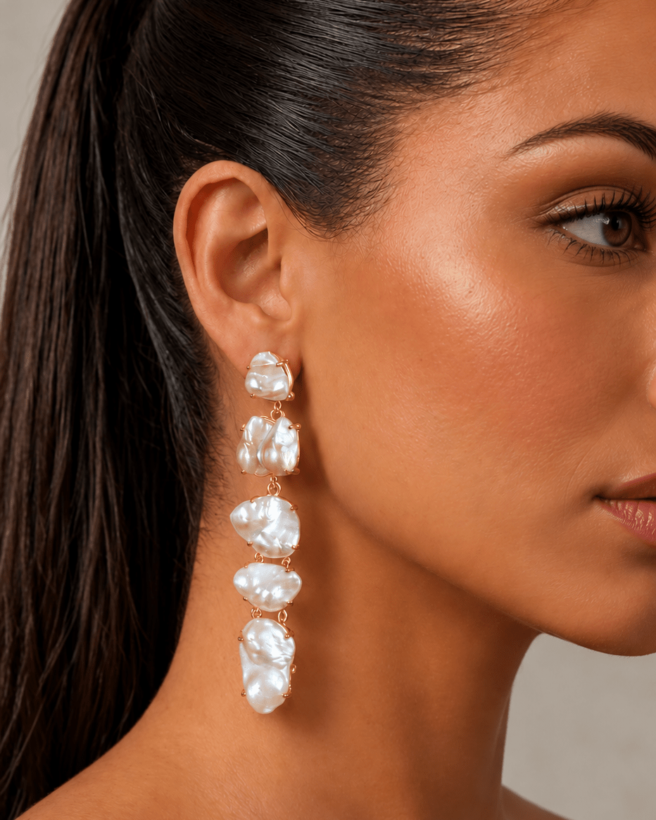 A close-up side profile of a woman wearing a long, dangling statement earring made of irregular white pearl-like beads set in gold-tone metal.