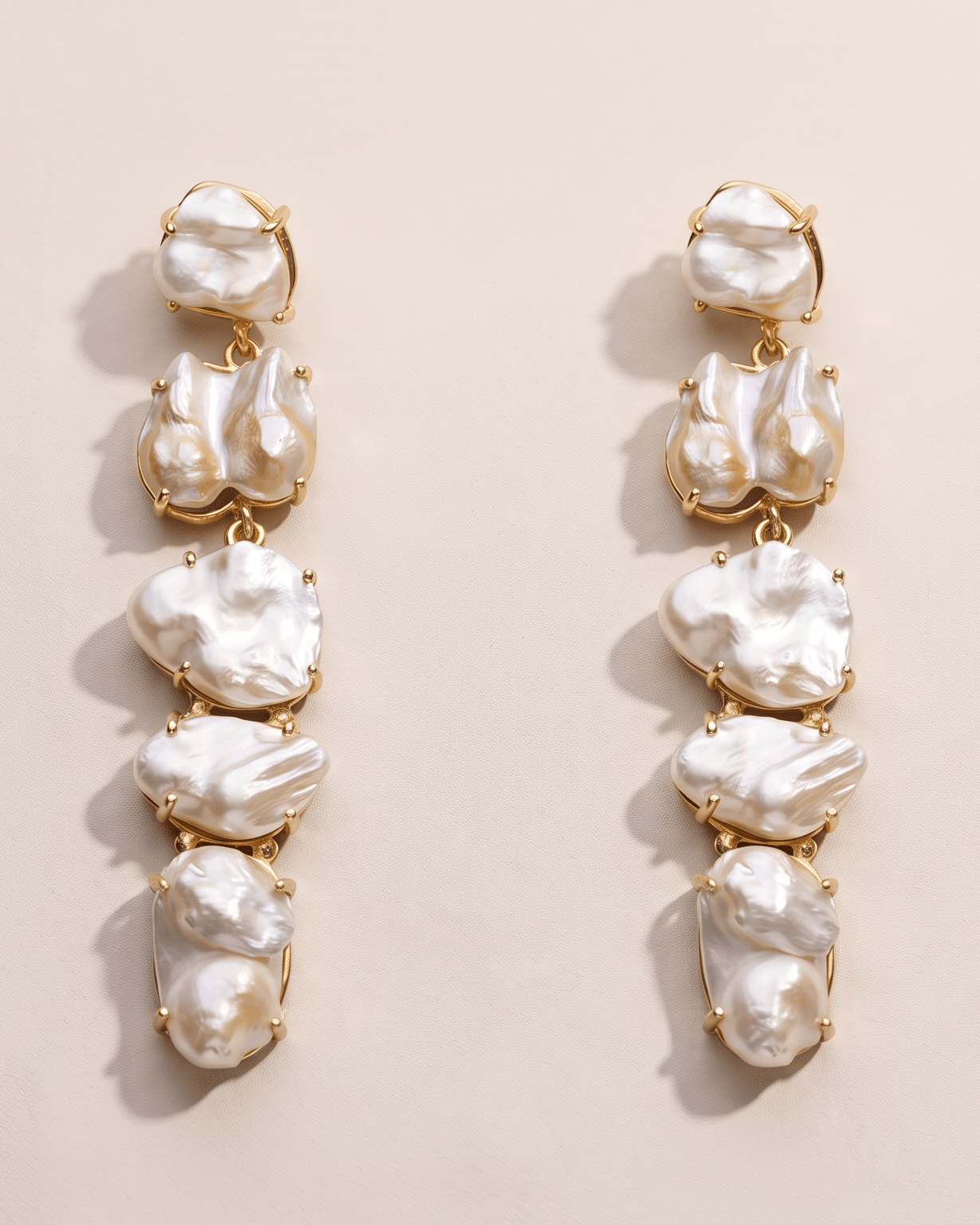 A pair of gold drop earrings featuring five vertical clusters of irregular, lustrous white baroque pearls, displayed on a warm beige background.