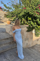 Woman in a light blue off-shoulder dress standing outdoors near stone steps and greenery.