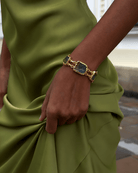 A close-up of a person wearing a gold-toned bracelet with rectangular blue-green stones, shown against a green satin dress and an outdoor blurred background.