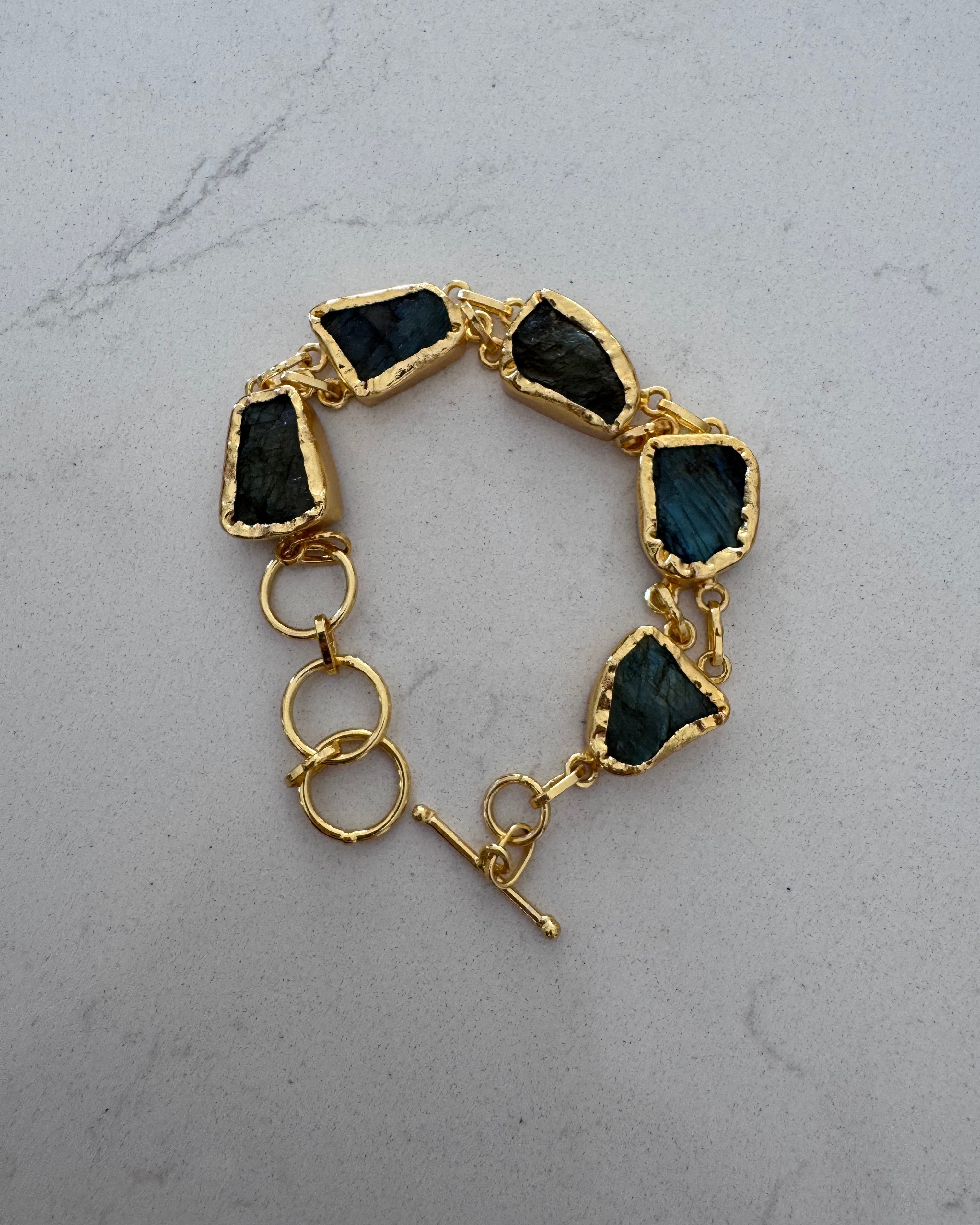 A gold-tone bracelet featuring five irregular dark blue-green stone settings and three interlocking rings, displayed on a light gray marble surface.