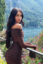 Woman in a brown dress standing by a scenic lakeside with mountains and flowers.